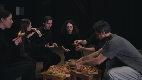 Confident-theater-actors-in-black-suits-pick-up-a-piece-of-pizza-and-give-lunches-during-a-break-during-a-rehearsal-in-a-theater-on-stage.-Close-up-of-a-confident-group-of-young-actors-eating-during-a-break-and-preparation-for-a-performance-in-the-theater