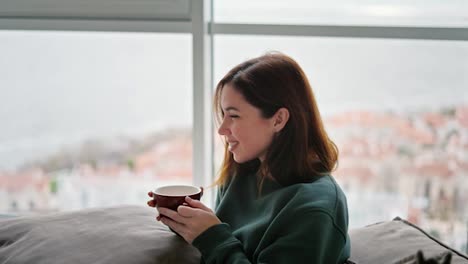 Side-view-of-a-happy-brunette-girl-with-long-hair-in-a-green-sweater-sits-on-a-modern-sofa-with-a-pillow-on-her-feet-and-the-girl-drinks-tea-from-a-brown-glass-in-a-modern-apartment-overlooking-the-sea