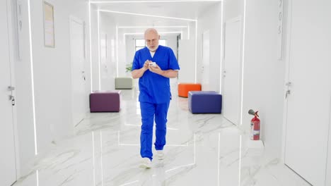 A-confident-male-doctor-in-glasses-with-a-gray-beard-in-a-blue-uniform-walks-along-a-bright-corridor-in-a-modern-clinic-and-types-on-a-white-smartphone