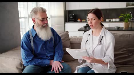 A-man-with-gray-hair-of-advanced-age-with-a-lush-gray-beard-in-a-blue-shirt-sits-on-a-modern-sofa-near-a-brunette-doctor-girl-in-a-white-coat-who-tells-him-about-methods-of-maintaining-health-in-a-modern-apartment