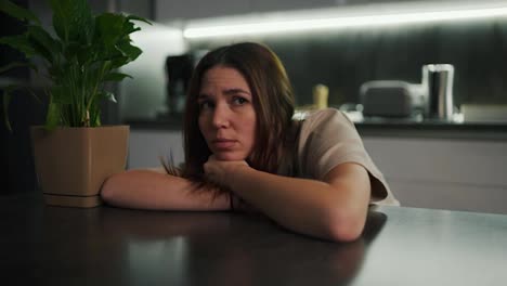 A-sad-and-tired-brunette-girl-in-a-beige-T-shirt-sits-leaning-on-a-black-dining-table-near-a-green-houseplant-in-a-modern-kitchen-in-the-evening