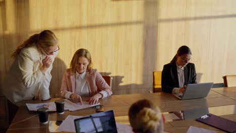 Side-view-of-a-group-of-businesswoman-in-business-suits-sitting-at-a-wooden-table-in-a-sunny-office-communicating-and-working-on-laptops