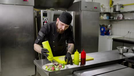 A-confident-male-cook-wearing-a-black-bandana-in-a-protective-uniform-and-wearing-black-rubber-gloves-uses-different-sauces-to-season-the-salad-and-prepare-it-for-serving-in-a-restaurant