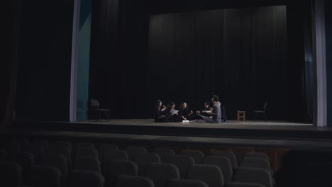 A-group-of-young-actors-in-black-uniforms-sit-on-stage-together-with-their-stage-director-communicate-and-analyze-the-script-while-preparing-for-a-performance-in-a-theater-with-a-black-curtain