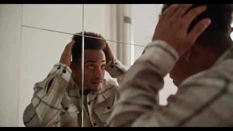 A-young-Black-skinned-brunette-man-with-stubble-in-a-cream-colored-checkered-shirt-preens-near-a-mirror-in-the-hallway-in-a-modern-apartment-with-light-walls-and-good-lighting