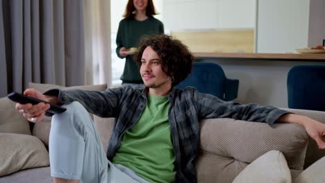 Happy-brunette-guy-with-curly-hair-in-a-gray-checkered-shirt-watches-TV-while-he-is-brought-sweet-snacks-by-his-brunette-girlfriend-in-a-modern-apartment-at-home