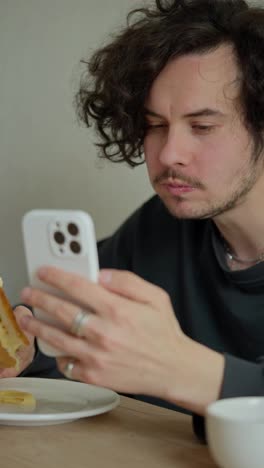 Vertical-video-of-a-brunette-guy-with-a-mustache-with-curly-hair-sitting-at-a-table-and-watching-videos-on-social-networks-while-eating-his-sandwich-in-the-morning