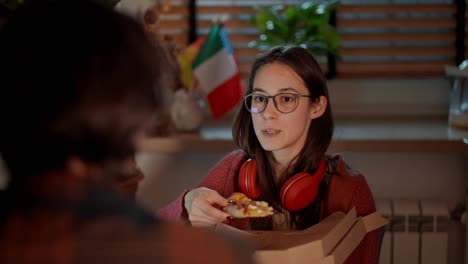 Over-the-shoulder-a-confident-brunette-girl-in-glasses-and-red-wireless-headphones-eats-pizza-during-her-snack-while-studying-foreign-languages-with-her-brunette-teacher-in-a-modern-apartment-in-the-kitchen-near-the-flags-of-Italy