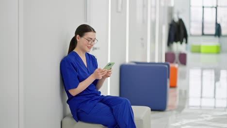 Confident-and-happy-girl-brunette-doctor-with-glasses-in-a-blue-uniform-sits-on-the-side-in-the-corridor-of-a-modern-clinic-and-types-on-a-green-smartphone