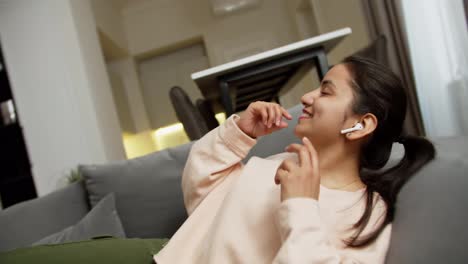 A-happy-brunette-Indian-girl-in-a-light-jacket-lies-down-on-the-sofa-while-listening-to-dynamic-music-in-white-wireless-headphones-in-a-modern-apartment