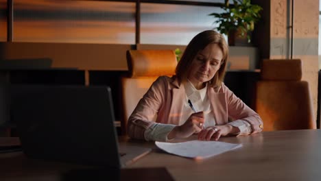 Concentrated-middle-aged-blonde-woman-in-a-pink-jacket-and-business-suit-sits-at-a-wooden-table-in-the-office-and-writes-down-her-ideas-on-paper-in-a-sunny-office-in-front-of-a-laptop