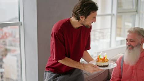 A-happy-young-man-with-a-brunette-with-stubble-brings-a-plate-with-a-small-cake-and-burning-candles-for-his-elderly-father-with-gray-hair-and-a-lush-beard-in-a-pink-shirt-during-his-congratulations-to-his-dad-on-his-birthday-in-a-modern-apartment