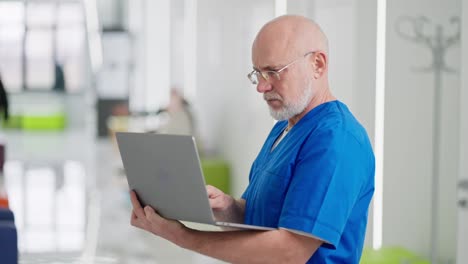 zoom-in-A-confident-elderly-man-with-glasses-a-doctor-in-a-blue-uniform-stands-in-the-middle-of-the-corridor-of-a-bright-clinic-and-types-on-his-gray-laptop