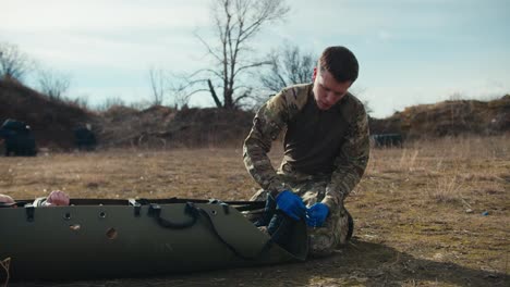 Side-view-of-a-confident-young-man-in-camouflage-uniform-and-blue-medical-gloves-tying-straps-on-a-special-military-stretcher-with-an-unconscious-soldier-in-the-steppe-during-combat-operations