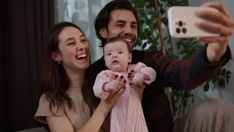 Happy-young-brunette-guy-with-stubble-together-with-his-brunette-wife-play-with-their-little-daughter-baby-in-a-pink-suit-and-take-a-family-selfie-photo-using-a-white-smartphone-in-a-modern-apartment