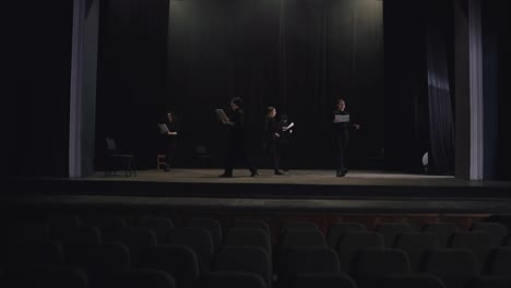 View-from-afar-a-confident-group-of-young-actors-in-black-suits-walk-around-the-stage-with-sheets-of-paper-in-their-minds-reading-their-text-and-script-of-the-play-and-preparing-for-their-performance-on-the-stage-of-a-theater-with-a-black-curtain