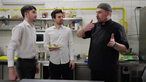 A-confident-male-chef-in-a-black-uniform-tells-two-young-guys-waiters-how-to-properly-take-out-dishes-and-work-in-the-kitchen-with-staff-in-a-restaurant