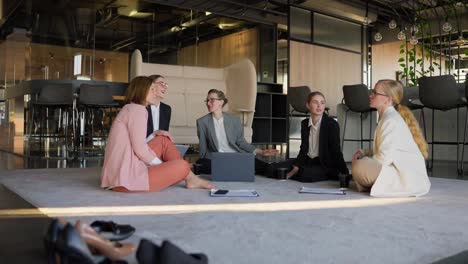 Zoom-in-approaching-a-group-of-confident-business-girls-in-business-uniforms-communicating-while-sitting-on-a-soft-carpet-in-a-modern-office