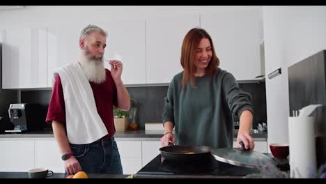 Ein-Glückliches-Brünettes-Mädchen-In-Einem-Grünen-Pullover-Bereitet-Das-Frühstück-Vor,-Während-Ihr-Vater,-Ein-älterer-Mann-Mit-Grauem-Haar-Und-üppigem-Bart,-In-Der-Küche-Einer-Modernen-Wohnung-Seine-Zähne-Putzt