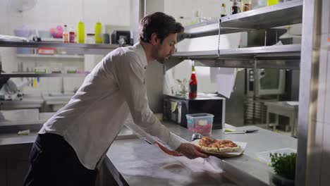 A-confident-brunette-male-waiter-in-a-shirt-takes-a-dish-and-delivers-it-to-the-hall-taking-it-from-the-serving-table-in-the-kitchen-in-a-restaurant.-Interaction-between-waiters-and-cooks-when-issuing-dishes-and-receiving-orders