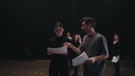 A-confident-young-brunette-man-the-stage-director-tells-how-to-play-his-role-correctly-and-say-the-text-of-a-brunette-girl-to-an-actress-in-a-black-suit-during-general-preparation-and-rehearsal-for-a-performance-in-a-theater-on-stage