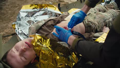 A-confident-male-medic-in-blue-medical-gloves-stops-the-blood-of-a-wounded-soldier-in-a-camouflage-uniform-who-is-lying-on-a-military-medical-stretcher-in-the-combat-area