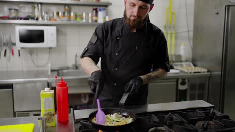 Confident-male-chef-with-a-beard-in-a-black-uniform-and-with-black-protective-gloves-fries-vegetables-in-a-frying-pan-adding-oil-in-a-modern-kitchen-in-a-restaurant