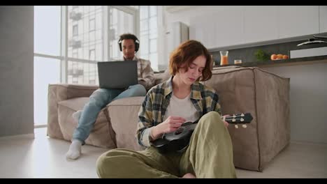 A-young-girl-with-a-bob-hairstyle-in-a-green-checkered-shirt-sits-near-the-sofa-on-the-floor-and-plays-the-ukulele-while-her-boyfriend-in-modern-Black-headphones-works-at-a-gray-laptop-in-a-modern-studio-apartment