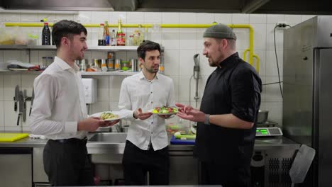 Side-view-of-a-professional-chef-in-a-black-uniform-communicating-with-two-waiters-in-white-shirts-holding-ready-made-dishes-about-solving-problems-in-the-kitchen-and-improving-dishes-and-their-serving-in-a-restaurant