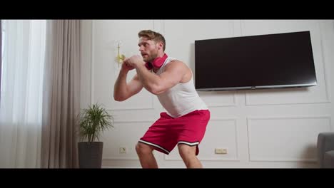 Confident-blond-man-with-stubble-in-a-white-T-shirt-and-red-shorts-doing-squats-while-doing-his-exercises-and-working-out-in-the-morning-at-home-in-a-modern-apartment