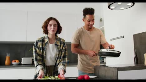 Portrait-of-a-happy-young-man-with-Black-skin-in-a-cream-T-shirt-and-a-young-adult-girl-with-a-bob-hairstyle-in-a-plaid-shirt-who-holds-in-her-hands-a-salad-and-a-fried-egg-while-preparing-breakfast-in-a-modern-kitchen