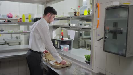 Side-view-of-a-confident-guy-waiter-in-a-white-shirt-putting-a-dish-into-a-square-cardboard-box-for-delivery-to-the-client.-Packing-food-in-a-restaurant-for-delivery