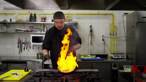 Confident-male-chef-in-a-black-uniform-adds-alcohol-to-a-frying-pan-with-fried-vegetables-and-makes-the-flambéed-technique-by-making-a-large-controlled-fire-on-the-stove-in-the-kitchen-in-a-restaurant