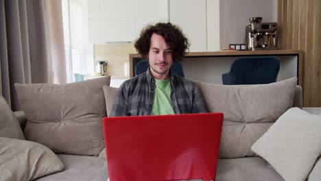 Focused-happy-brunette-guy-with-curly-hair-in-a-gray-checkered-shirt-sits-on-the-sofa-and-works-at-his-red-laptop-in-a-modern-studio-apartment