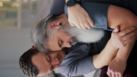 Vertical-video-of-a-sad-man-looking-for-support-and-leaning-on-his-brunette-boyfriend-with-stubble-in-a-gray-T-shirt-while-sitting-on-the-sofa-in-a-modern-apartment.-Elderly-man-leans-on-his-boyfriend-during-sad-mood-LGBT-couple