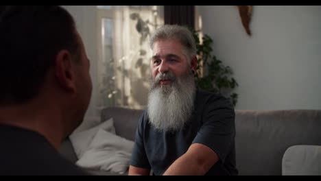 close-up-Over-the-shoulder-of-a-happy-man-with-gray-hair-and-a-lush-gray-beard-hugging-his-brunette-boyfriend-in-a-gray-T-shirt-in-a-modern-apartment-on-the-sofa-in-the-evening