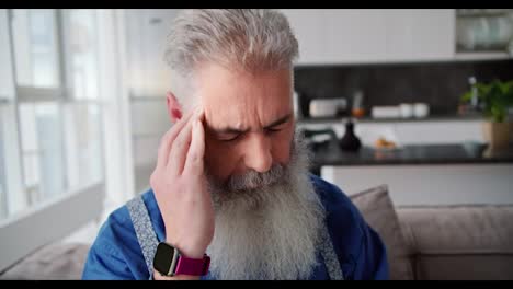 Close-up-of-an-elderly-man-with-gray-hair-and-a-full-beard-in-a-blue-shirt-massages-his-temple-during-a-headache-while-sitting-on-a-sofa-in-a-modern-apartment