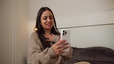 A-happy-brunette-Indian-girl-in-a-gray-sweater-talks-about-her-impressions-while-blogging-for-her-viewers-in-a-modern-apartment-in-the-bedroom-while-sitting-on-the-bed