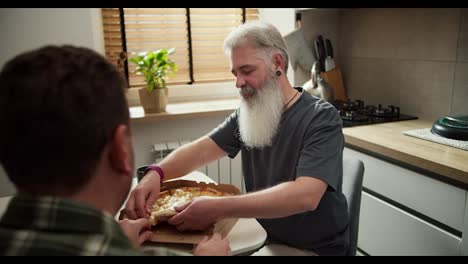 Happy-man-with-gray-hair-color-and-lush-white-beard-with-a-gray-T-shirt-eats-pizza-with-his-brunette-boyfriend-in-a-plaid-shirt-in-the-kitchen-from-a-cardboard-delivery-box