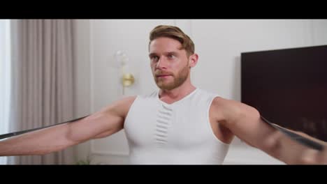 A-confident-blond-man-with-an-athletic-build-and-stubble-in-a-white-T-shirt-makes-swings-with-his-arms-and-does-morning-exercises-using-a-special-elastic-band-in-a-modern-apartment-in-the-morning-at-home