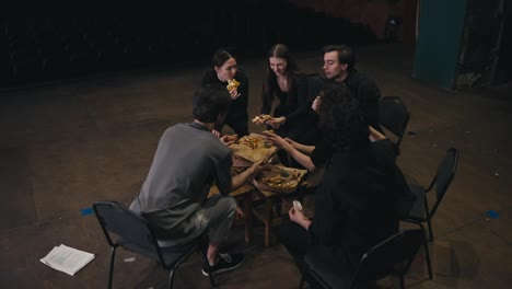 A-confident-small-group-of-actors-in-black-suits-sits-in-a-semicircle-and-is-given-pizza-for-lunch-while-preparing-for-a-performance-along-with-their-stage-director-on-the-theater-stage