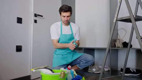 Selbstbewusster-Brünetter-Mann-Mit-Dreitagebart-In-Weißem-T-Shirt-Und-Blauer-Schürze-Als-Hausmeister,-Der-Auf-Dem-Boden-Zwischen-Reinigungswerkzeugen-In-Einem-Modernen-Apartment-Sitzt