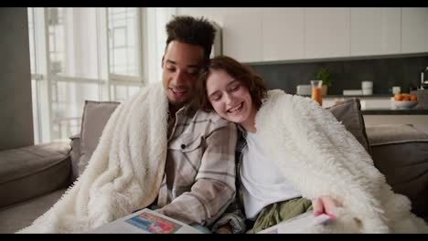 Happy-young-man-with-Black-skin-color-in-a-plaid-shirt-sits-with-his-girlfriend-on-a-modern-gray-sofa-wrapped-in-a-white-wool-blanket.-They-read-a-book-and-stories-in-a-modern-apartment-while-spending-time-together