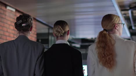 Rear-view-of-a-group-of-confident-businesswomen-in-business-clothes-walking-along-the-corridor-in-a-modern-office.-Confident-girls-in-business-clothes-walk-along-the-office-corridor-together