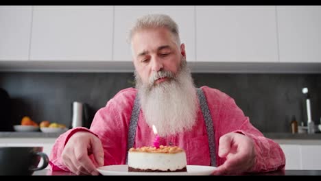 Un-Hombre-Triste-Y-Solitario,-De-Cabello-Gris-Y-Barba-Exuberante-Con-Una-Camisa-Rosa,-Mira-Un-Pastel-En-El-Que-Hay-Una-Vela-Encendida-Durante-Su-Cumpleaños-Y-La-Apaga-En-Un-Apartamento-Moderno.