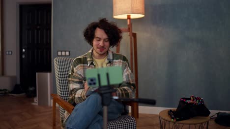 Happy-brunette-guy-with-curly-hair-in-a-checkered-shirt-talks-to-his-blog-subscribers-using-a-green-smartphone-and-writes-down-important-things-at-home-in-an-atmospheric-room