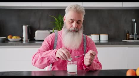 A-serious-elderly-man-with-gray-hair-and-a-lush-beard-in-a-pink-shirt-pours-white-medicine-into-a-glass-and-stirs-it-with-water-using-a-spoon-in-a-modern-kitchen