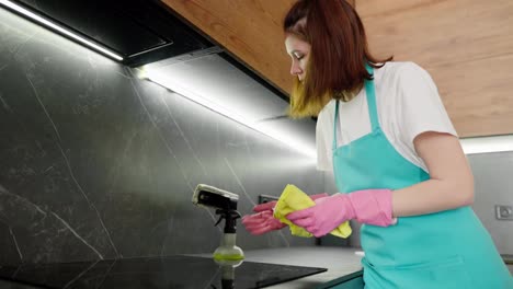 A-brunette-cleaning-lady-girl-in-a-white-T-shirt-and-a-blue-apron-uses-detergent-and-a-rag-to-wash-a-black-electronic-stove-in-the-kitchen-in-a-modern-apartment-on-a-cleaning-call