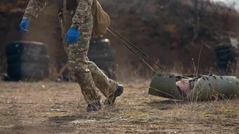 Close-up-a-man-lies-unconscious-on-a-green-army-carrier-for-the-wounded-while-a-medical-army-soldier-drags-him-along-the-steppe-at-an-army-training-ground