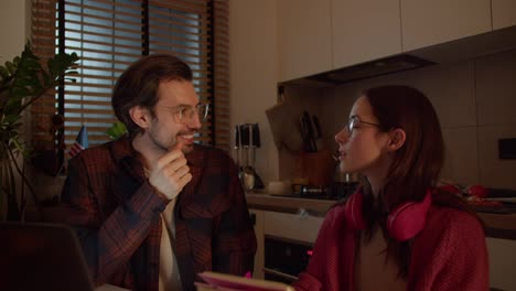 A-confident-young-brunette-girl-with-glasses-in-a-red-sweater-and-wireless-headphones-studies-foreign-languages-together-with-her-brunette-male-teacher-who-teaches-the-girl-the-correct-pronunciation-in-a-modern-apartment-in-the-kitchen